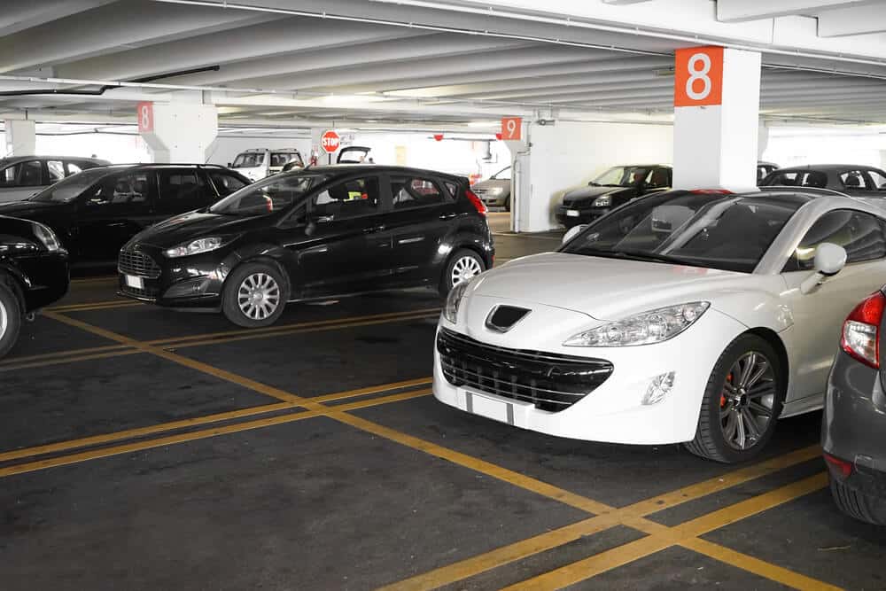 Cars parked in a multi-level parking garage with numbered sections; a white car and a black car are prominent in the foreground, marked section 8 on white pillars. The efficient parking management system ensures smooth navigation through this commercial parking facility.