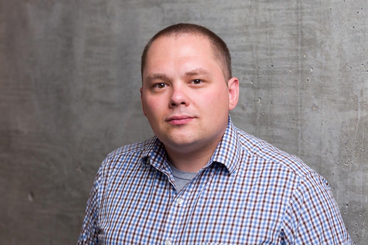 A man in a checkered shirt stands in front of a textured concrete wall, looking directly at the camera, perhaps ready to discuss efficient parking lot management.