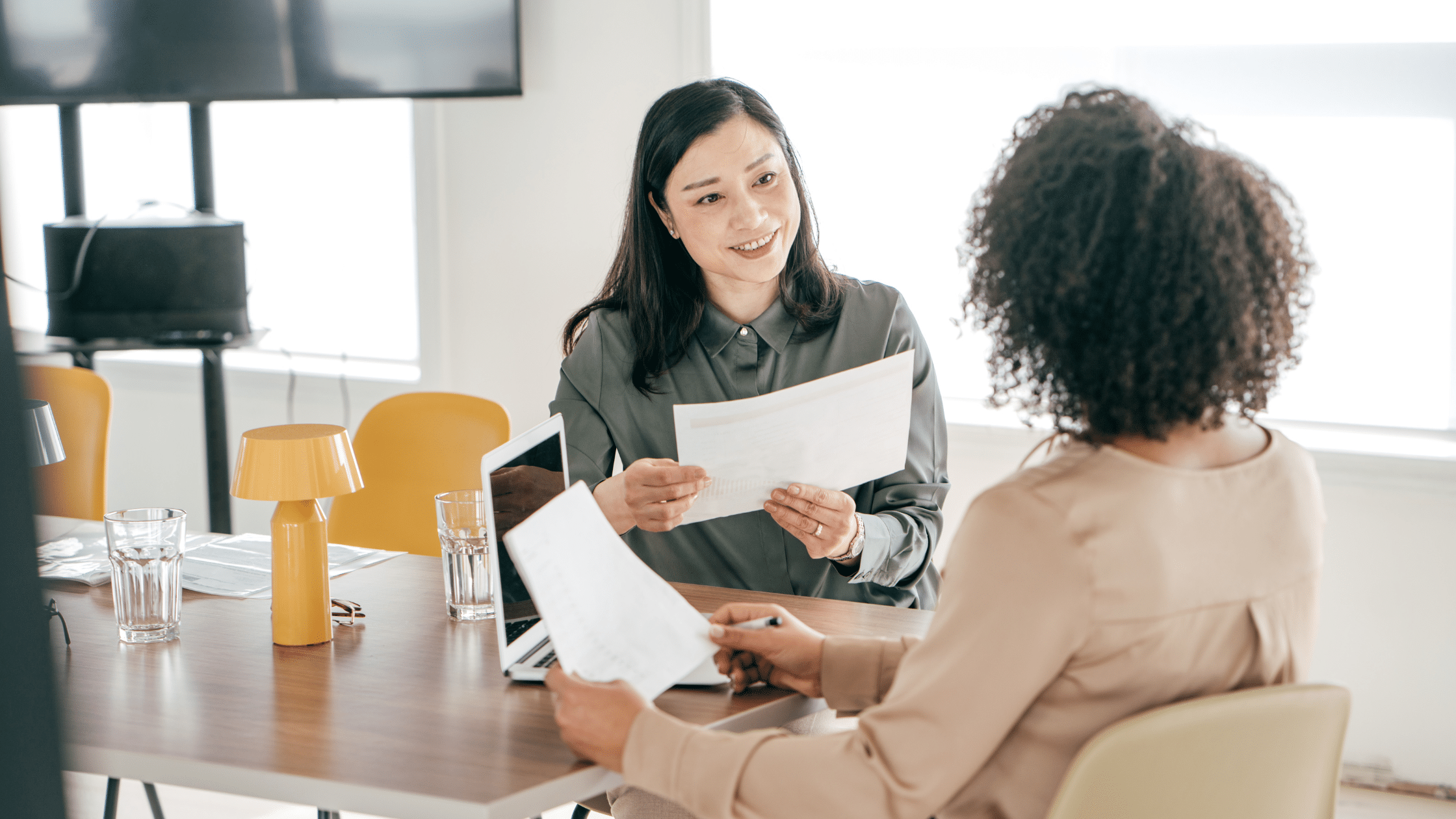 Two women are sitting at a table in an office, discussing documents related to parking lot management. One woman is handing a paper to the other, who holds another document as well. A laptop with Wayleadr software and a yellow lamp are on the table.