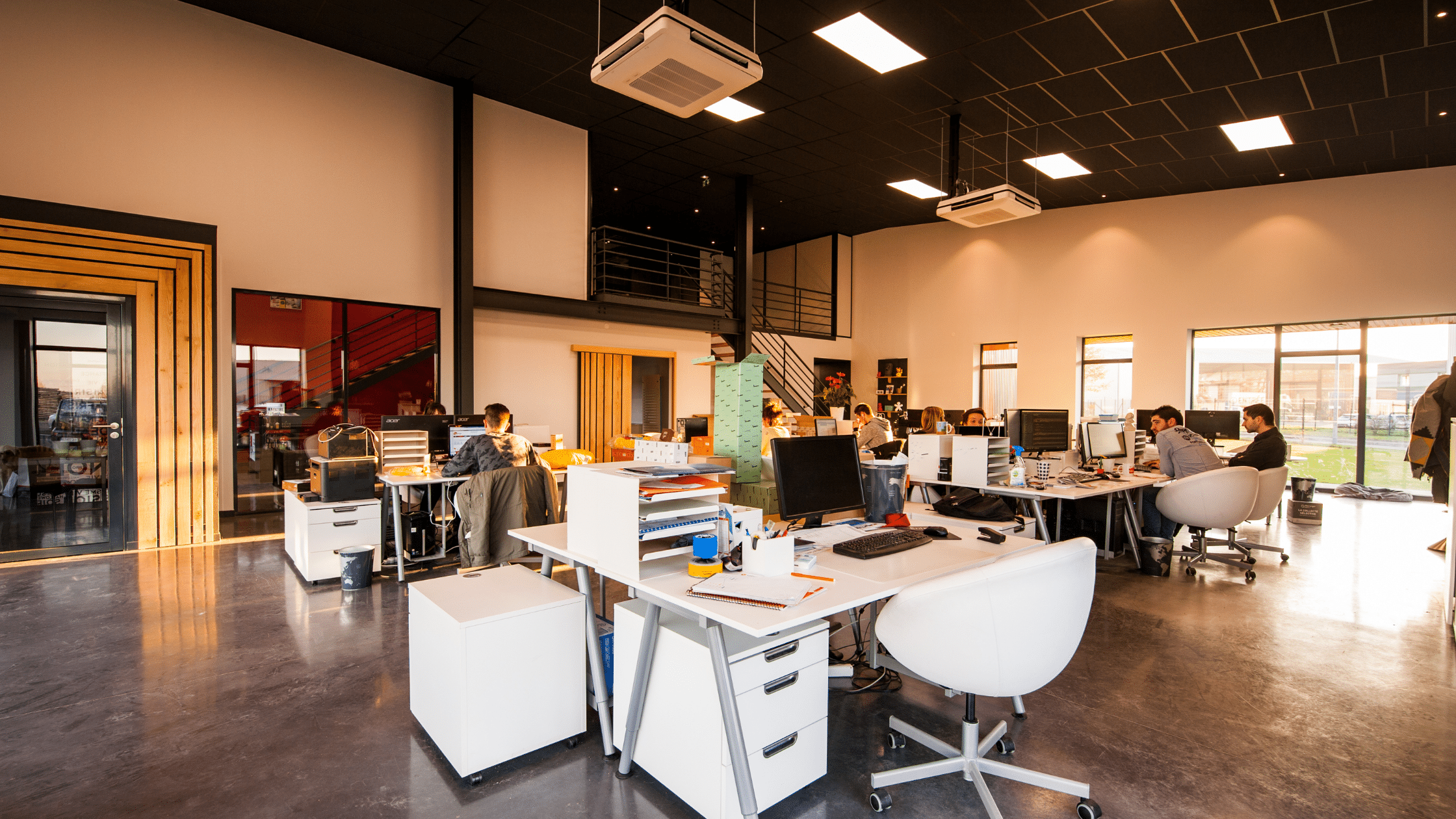 Open-plan office with several desks, computers, and chairs. People are working at their stations while managing visitor parking through Wayleadr. Large windows allow natural light. An upper loft area is visible in the background.