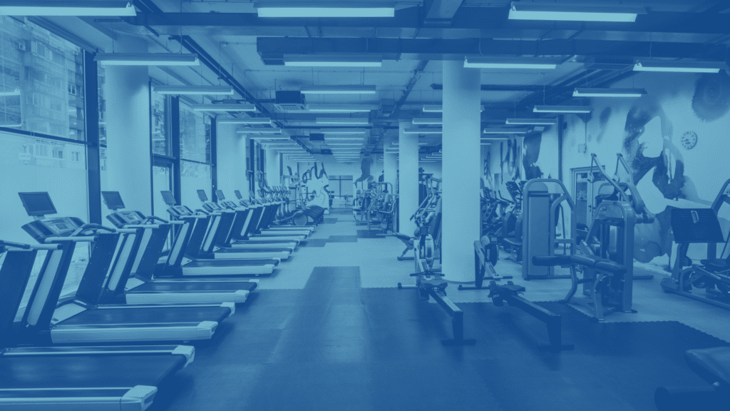 A spacious indoor gym with multiple treadmills, exercise bikes, and strength training equipment lined up neatly under bright fluorescent lighting.