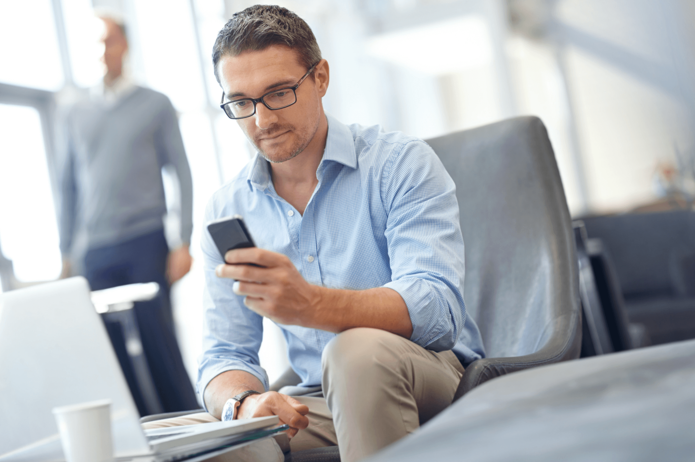 Man in glasses using a smartphone while sitting on a chair in a bright office space. A laptop and a coffee cup are on the table in front of him. Another person stands in the background.