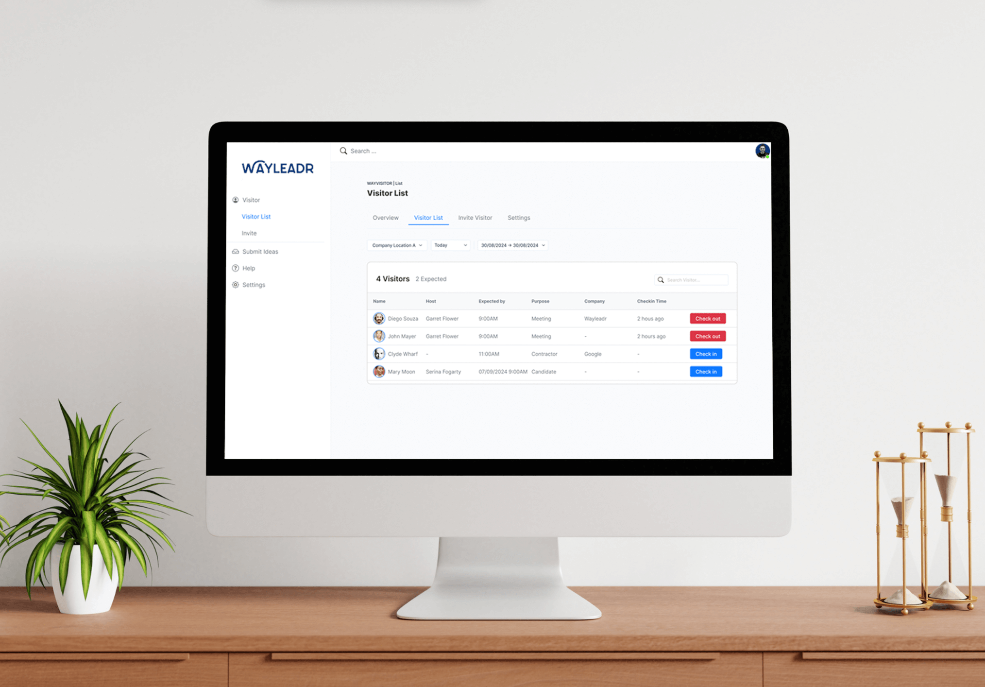 Computer monitor displaying a visitor list interface with check-in and check-out options. A small plant and an hourglass are on the wooden desk.