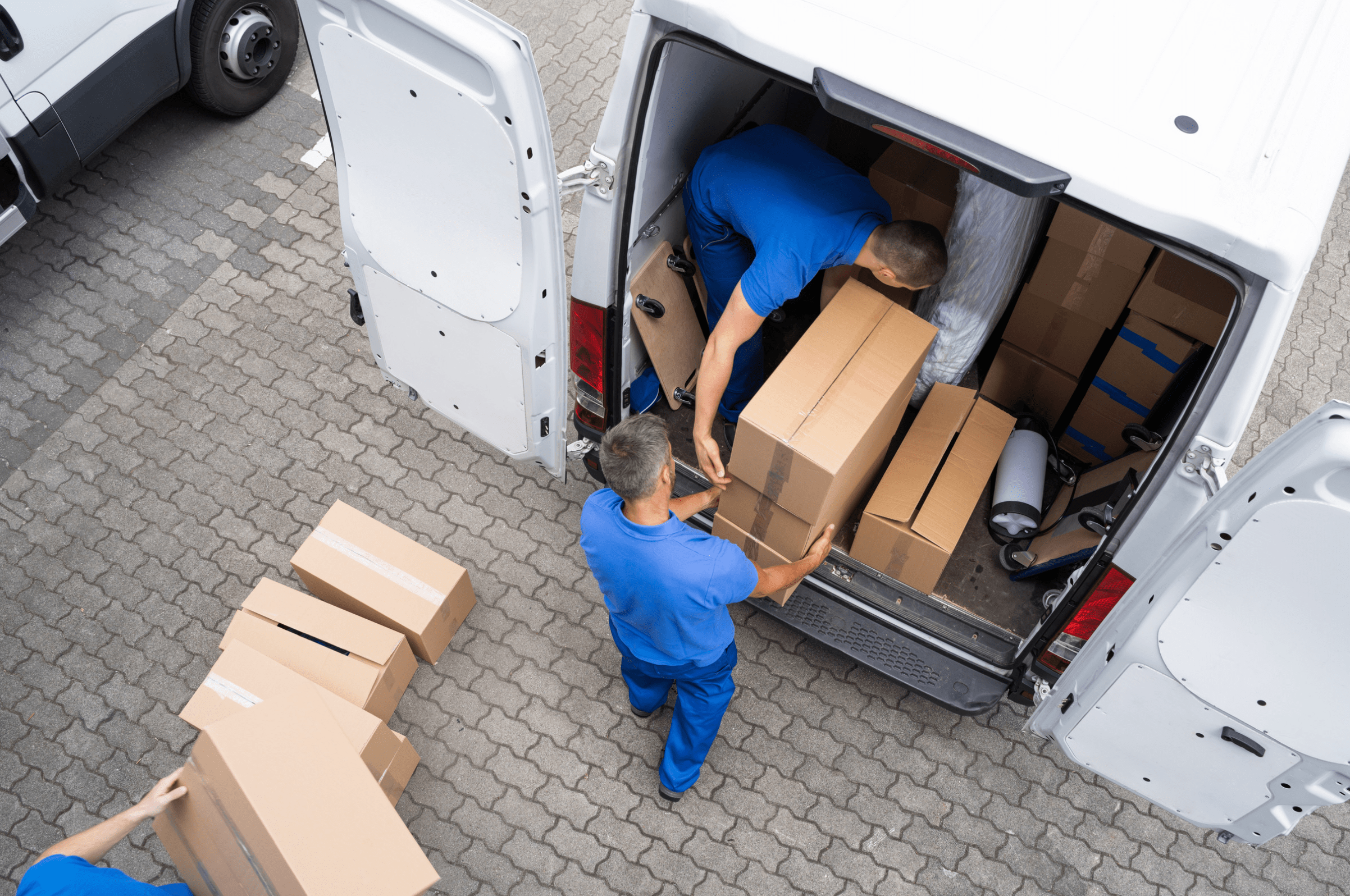 Workers in blue uniforms loading cardboard boxes into the back of a white delivery van on a paved surface.