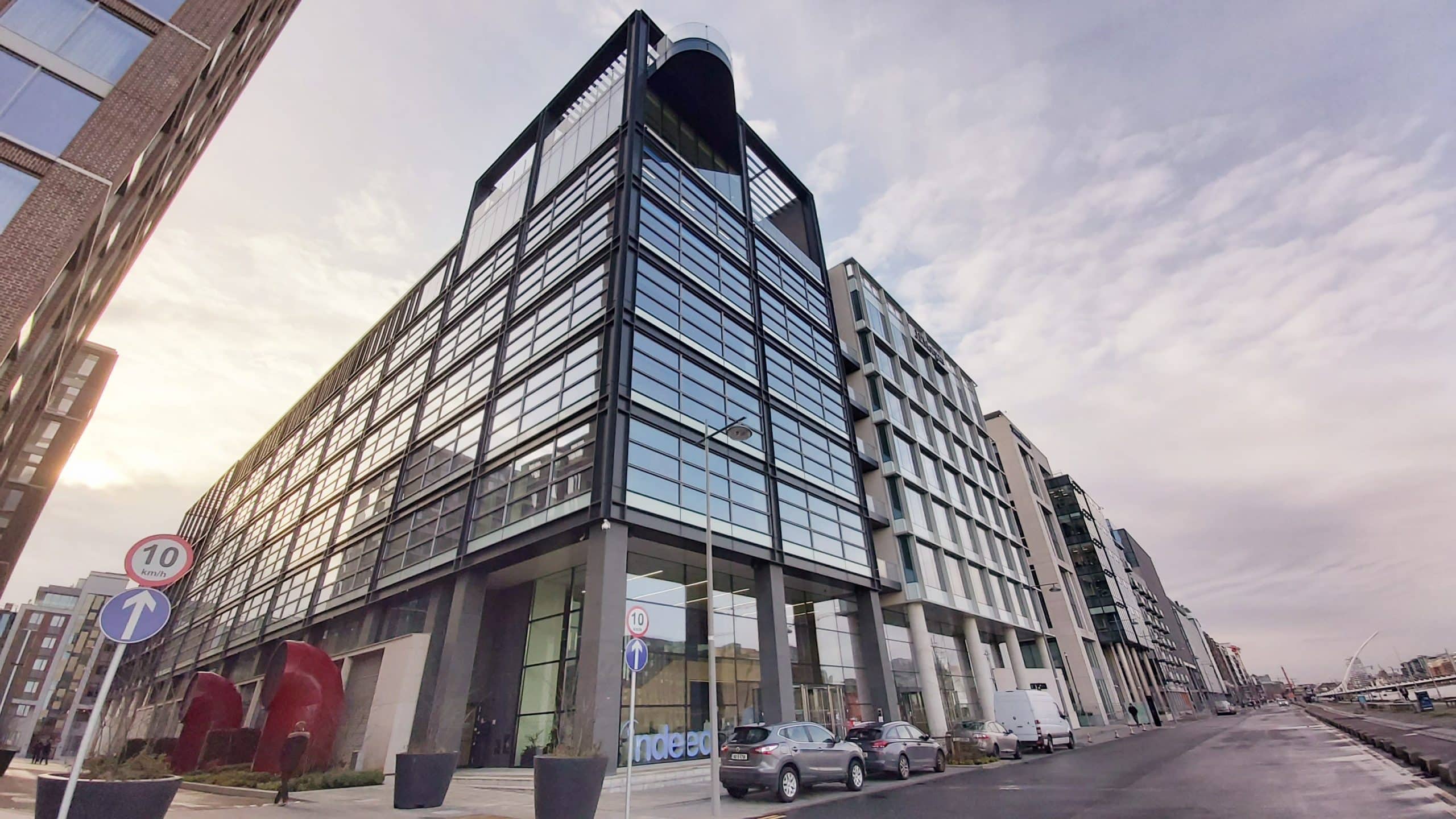 A modern, glass-fronted office building with cars parked along the street, flanked by a 10 km/h speed limit sign and a sunset in the background.