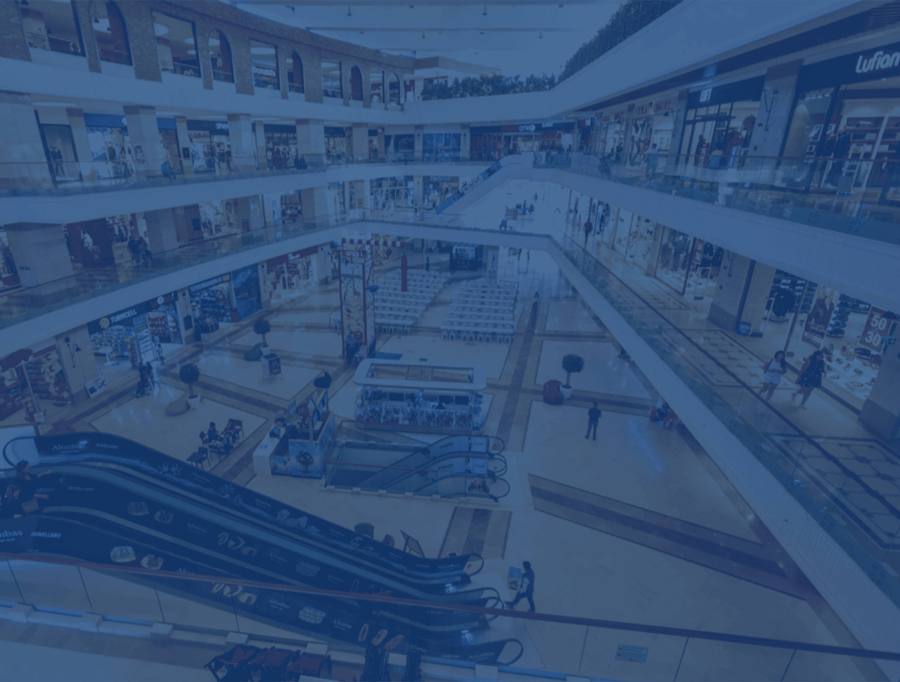 A spacious, multi-level shopping mall with escalators, stores on each level, and a central atrium view, enhanced with a blue filter.